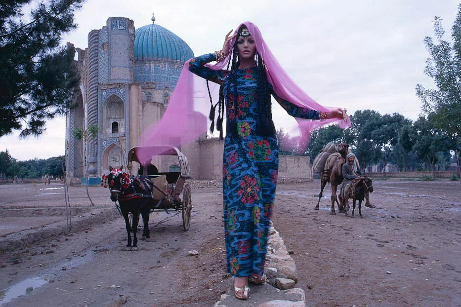 Model in a flowing blue dress walking past a horse-drawn carriage and blue-domed mosque.